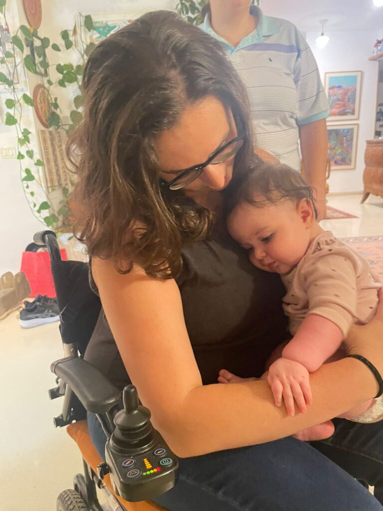 A woman in a wheelchair cuddles a baby, exuding warmth and emotional support in a cozy room filled with plants.